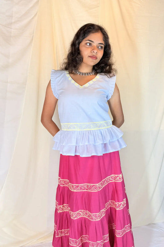 Woman wearing a light blue top and pink skirt with white patterns against a beige curtain background