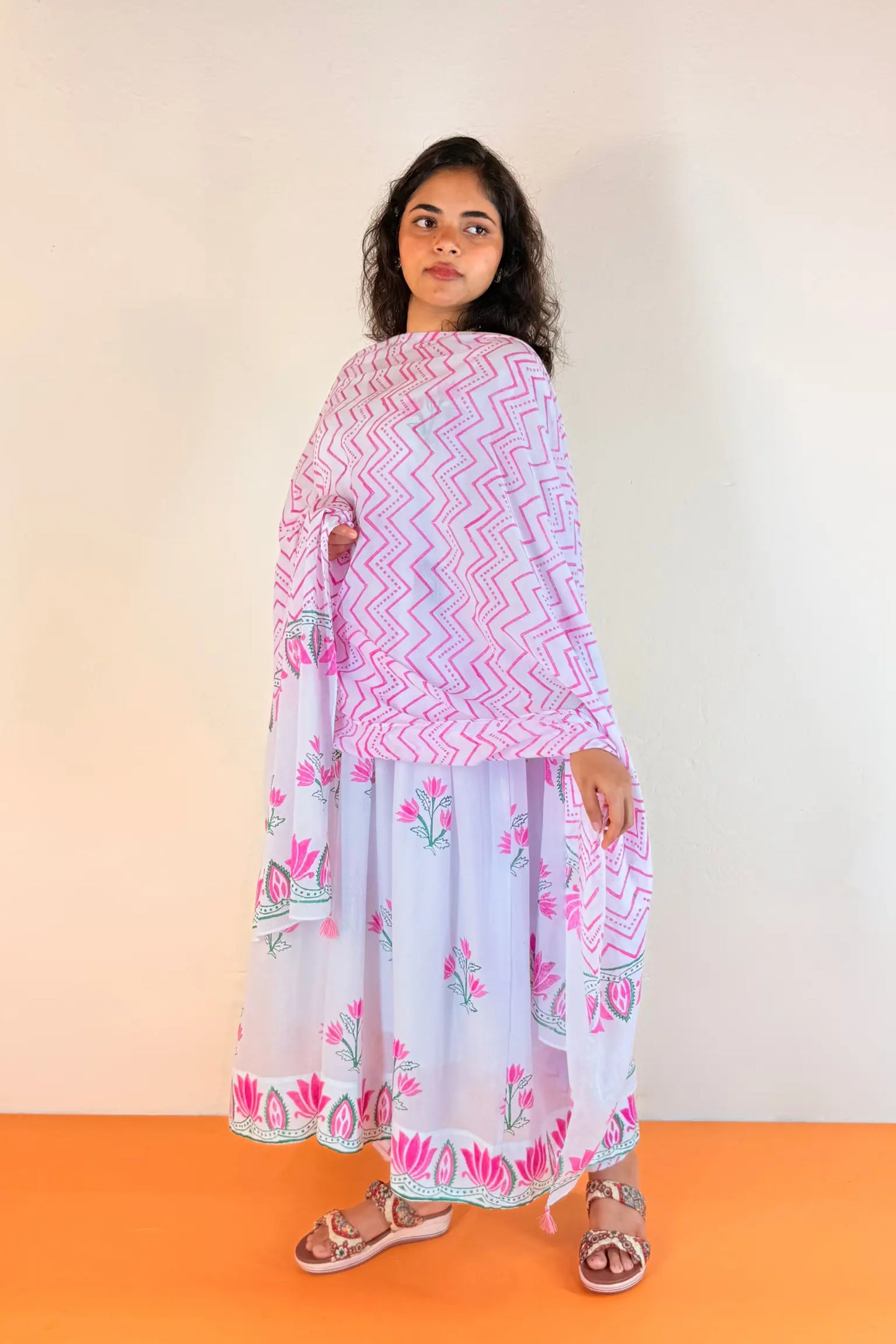 A woman modeling a traditional Indian outfit consisting of a white kurta with pink and blue chevron patterns and a matching printed dupatta, paired with white wide-leg pants and brown sandals, set against a plain off-white wall and orange floor.