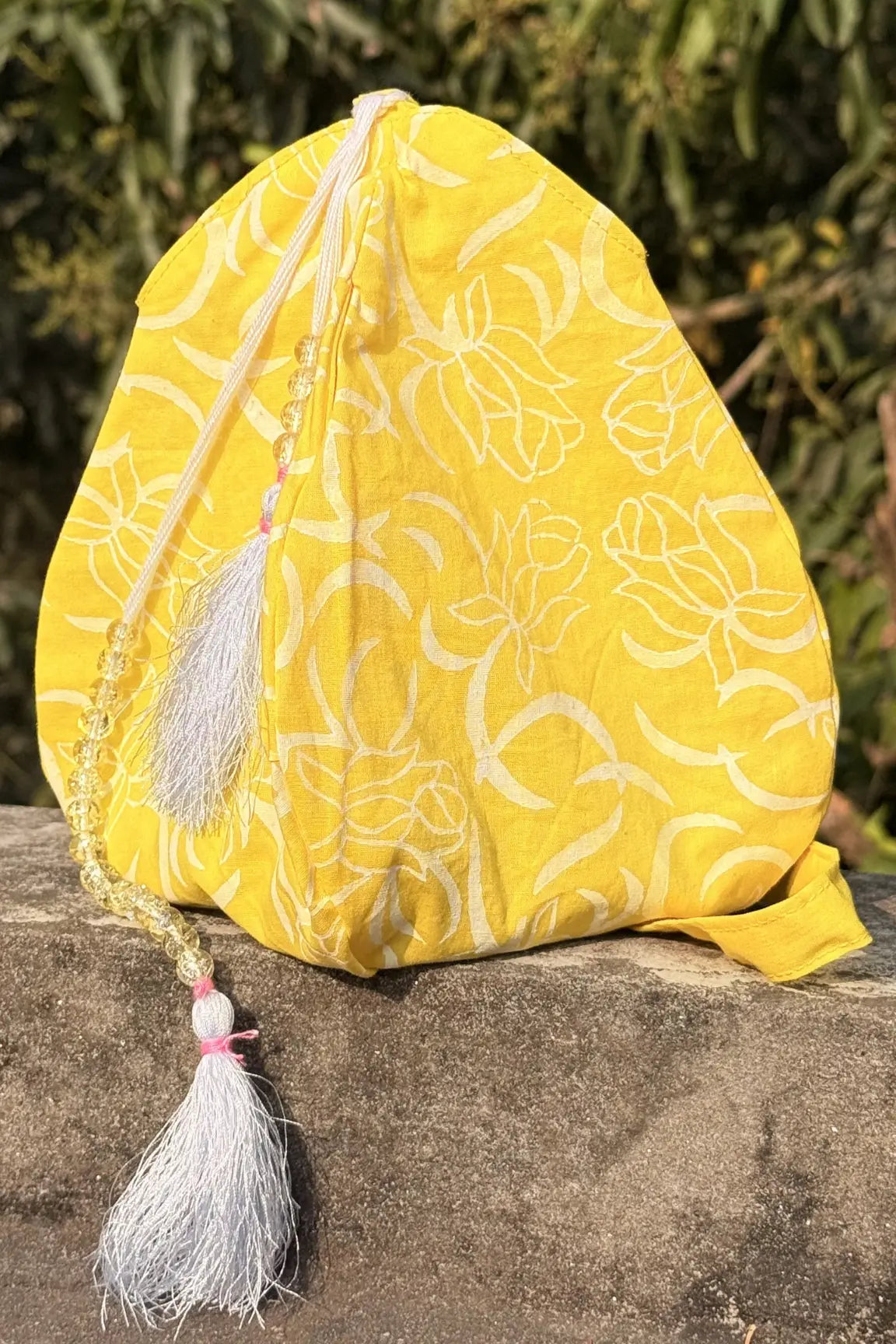 Yellow fabric pouch with floral patterns and tassels on a stone surface with green foliage in the background