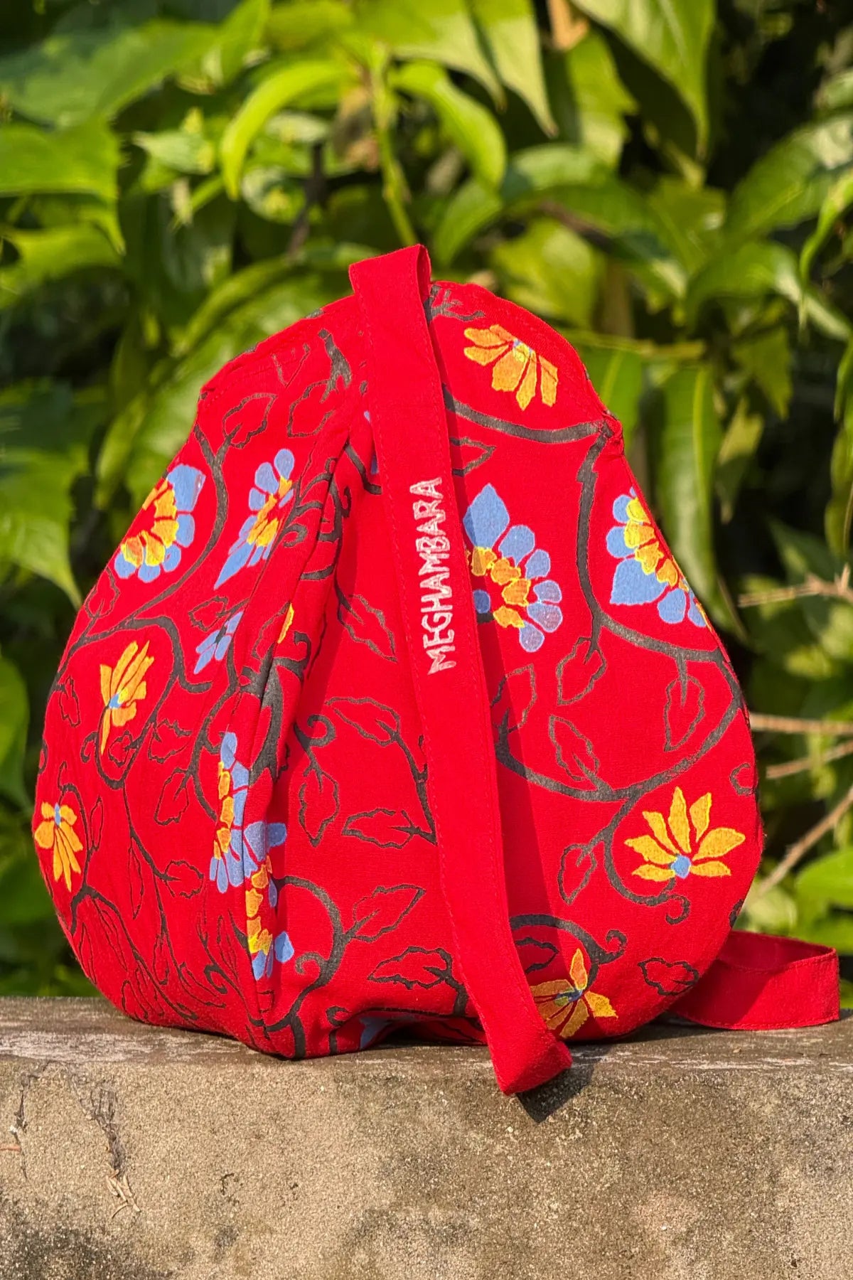 Hand block printed red cotton japa mala bag with multicolor floral motif and Meghambara branding strap, photographed outdoors