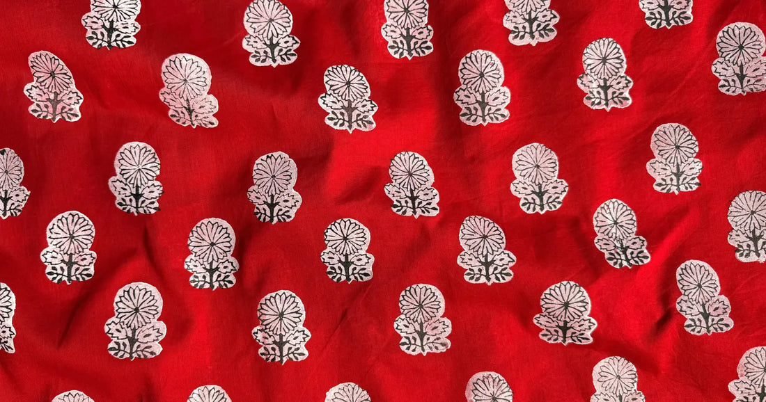 Red fabric featuring a repeating pattern of small floral block prints in white and black, showcasing the texture of handcrafted textile.