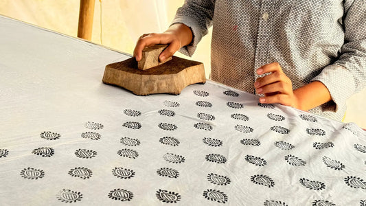 Hands of an artisan block printing fabric by hand, pressing a wooden block onto cotton cloth with repeated motifs, reflecting slow and thoughtful craftsmanship.