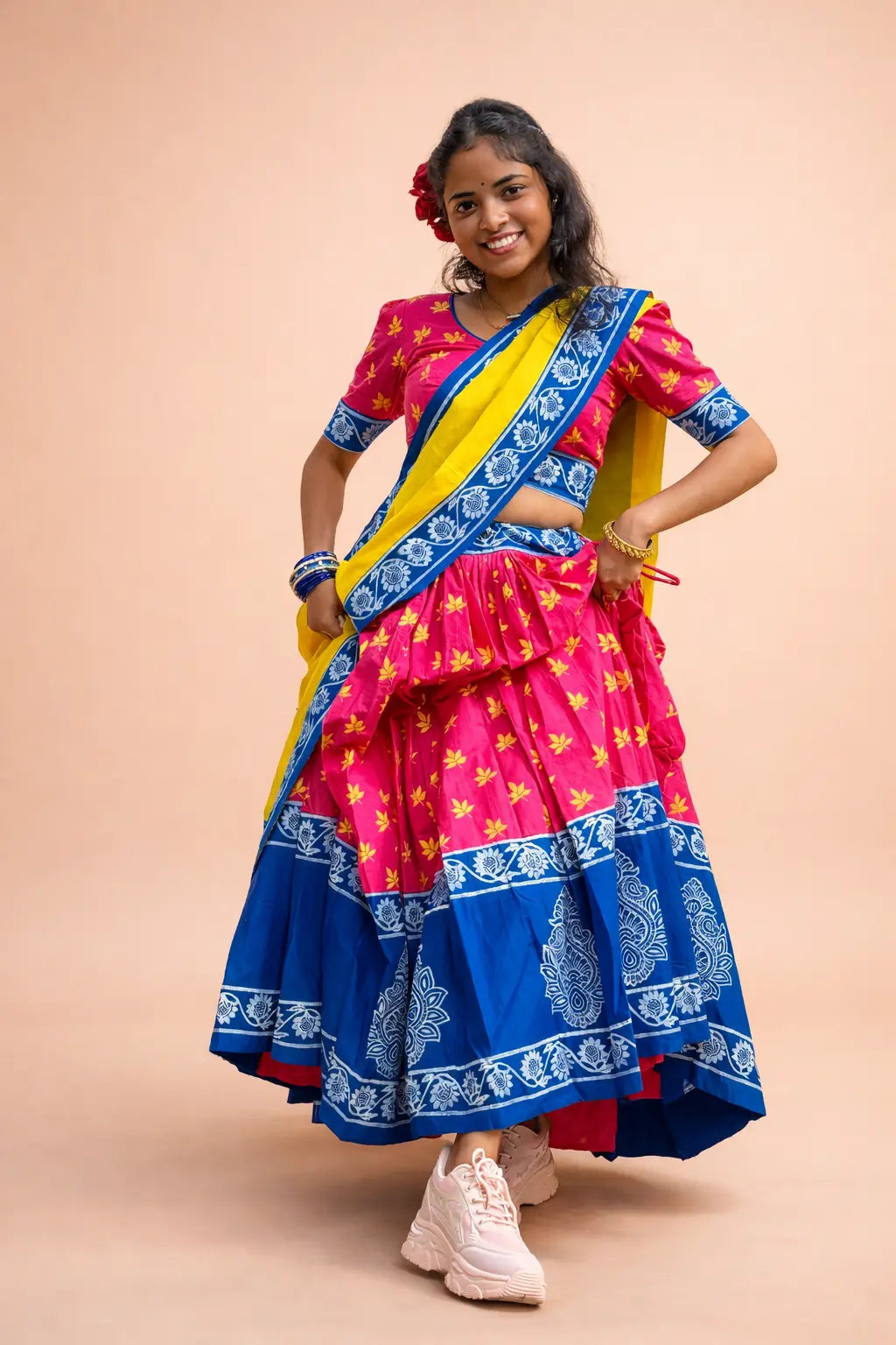 Smiling woman wearing a vibrant pink and blue hand block printed cotton festive ethnic dress with yellow dupatta, posing in a studio against a soft peach background.