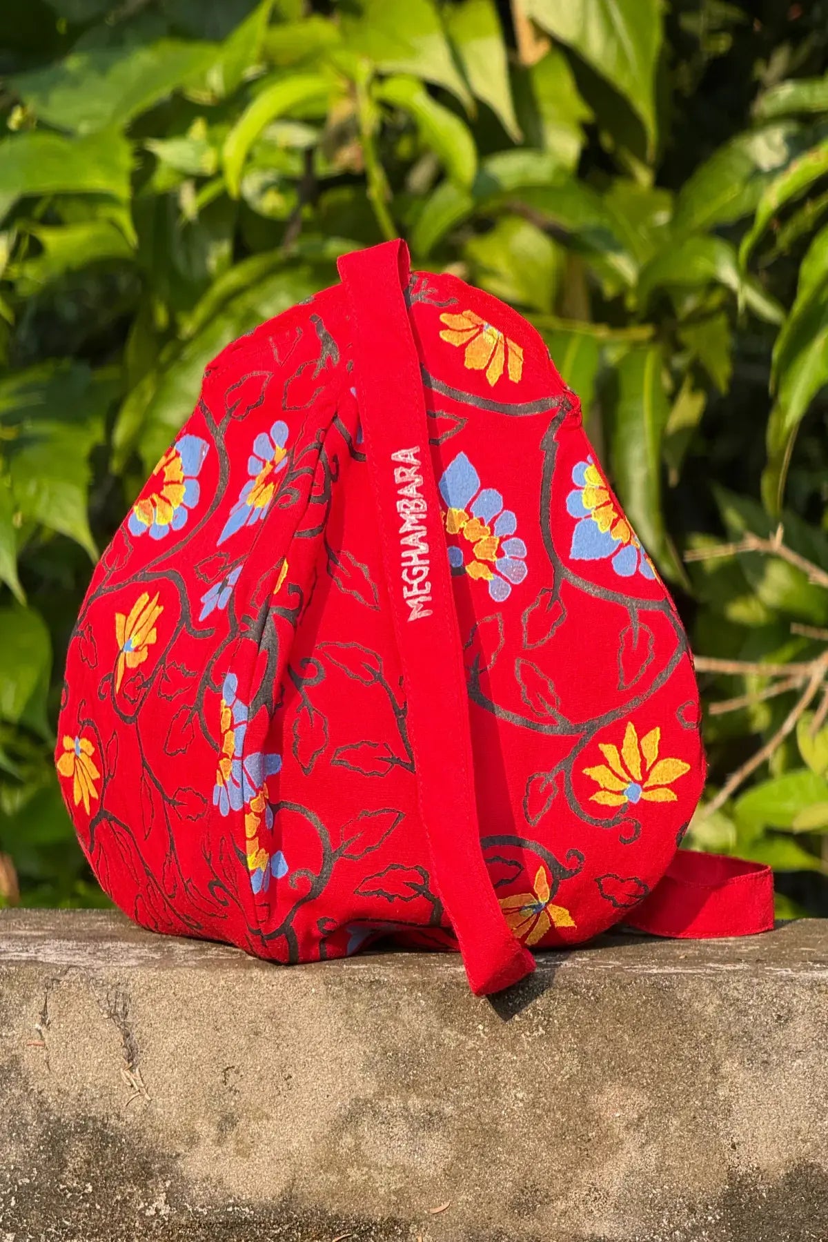 Red hand block printed fabric bag with floral motifs and Meghambara brand label, placed outdoors on a stone surface with green leaves in natural sunlight.