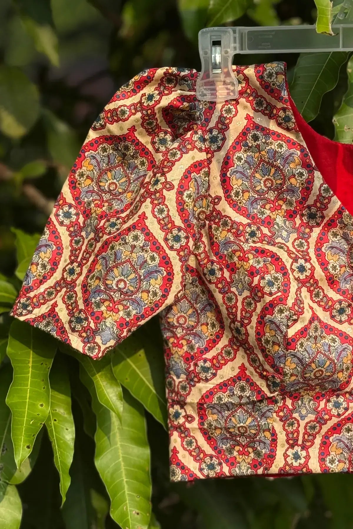 Hand block printed cotton blouse in beige and red floral motif, hanging on a clip outdoors with green leaves in natural sunlight.