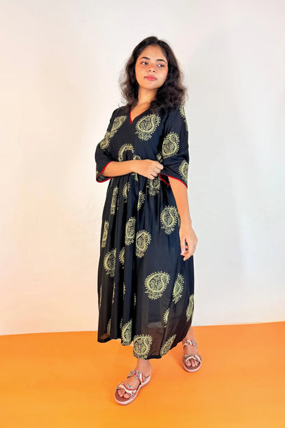 A woman modeling a black midi kaftan dress with green paisley patterns, standing against a plain white wall with an orange floor.