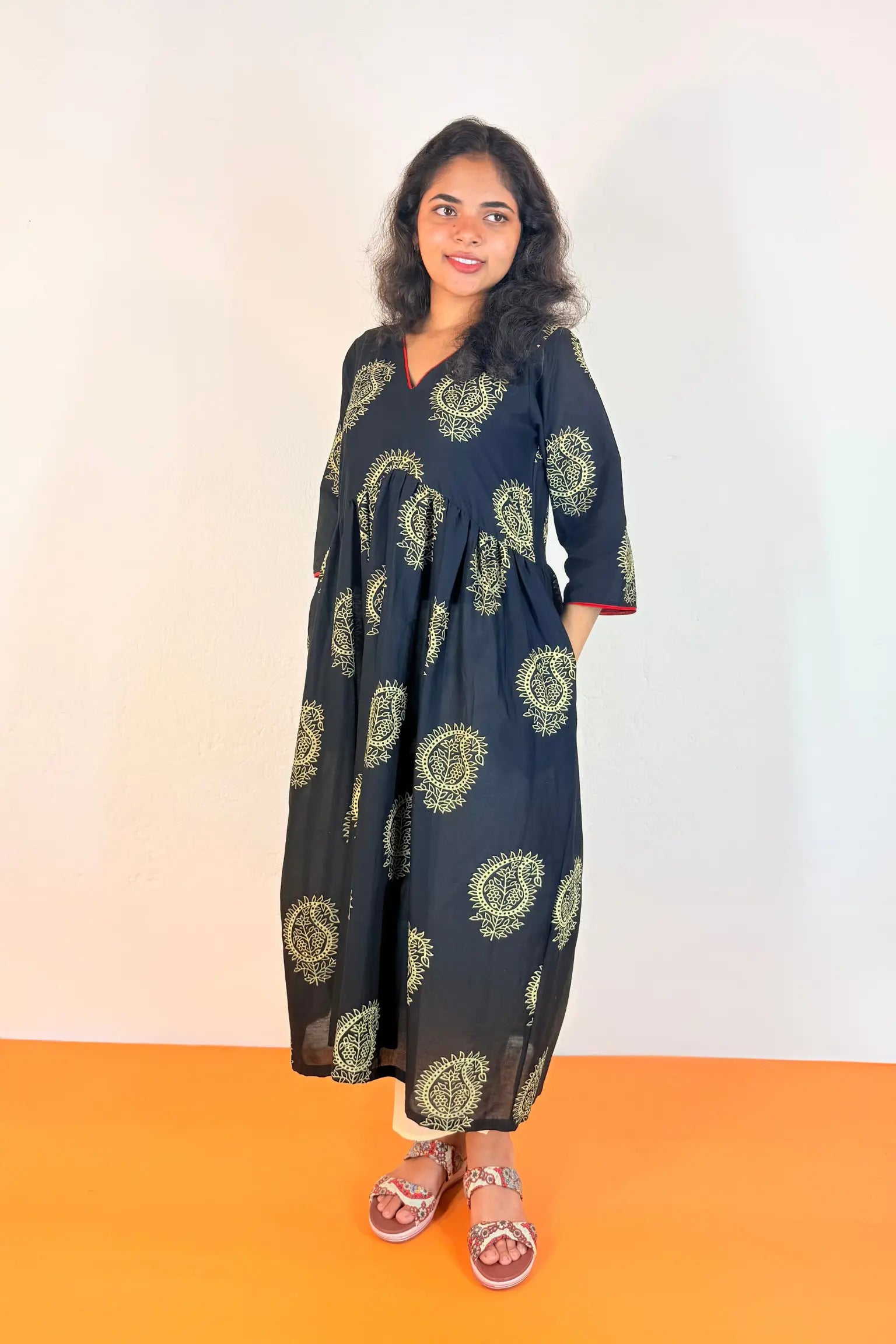 A woman modeling a light blue and white printed traditional outfit with a matching dupatta, standing against a plain off-white wall and orange floor.