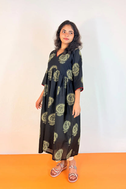 A woman modeling a black cotton kaftan dress with a beige and green paisley pattern, standing against a plain white wall with an orange floor.