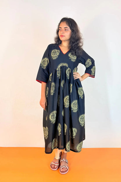 A woman modeling a black kaftan dress with green paisley block prints, standing against a plain white wall with an orange floor.