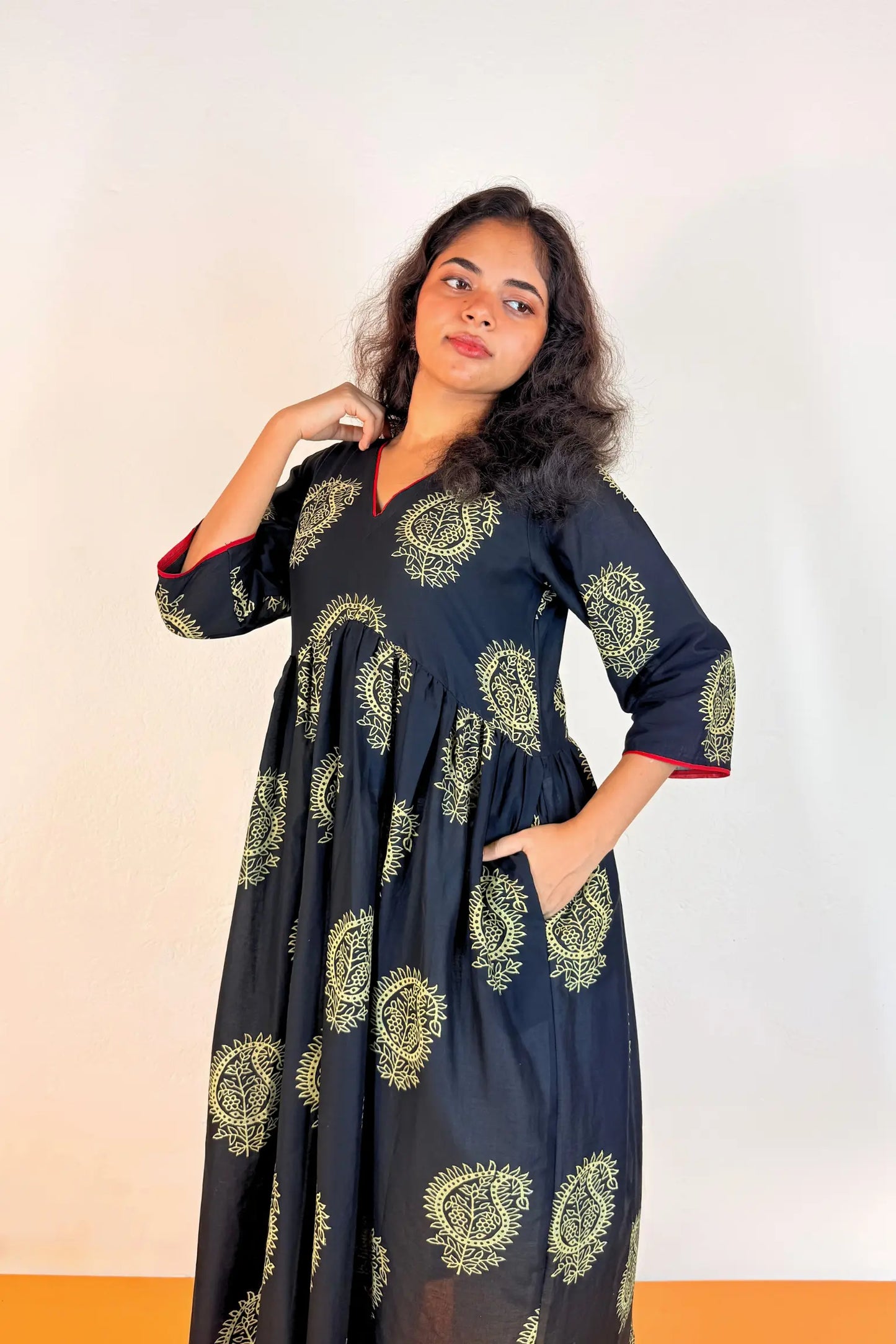A woman modeling a long black dress with beige paisley patterns and red lining on the sleeves, standing against a plain light background.