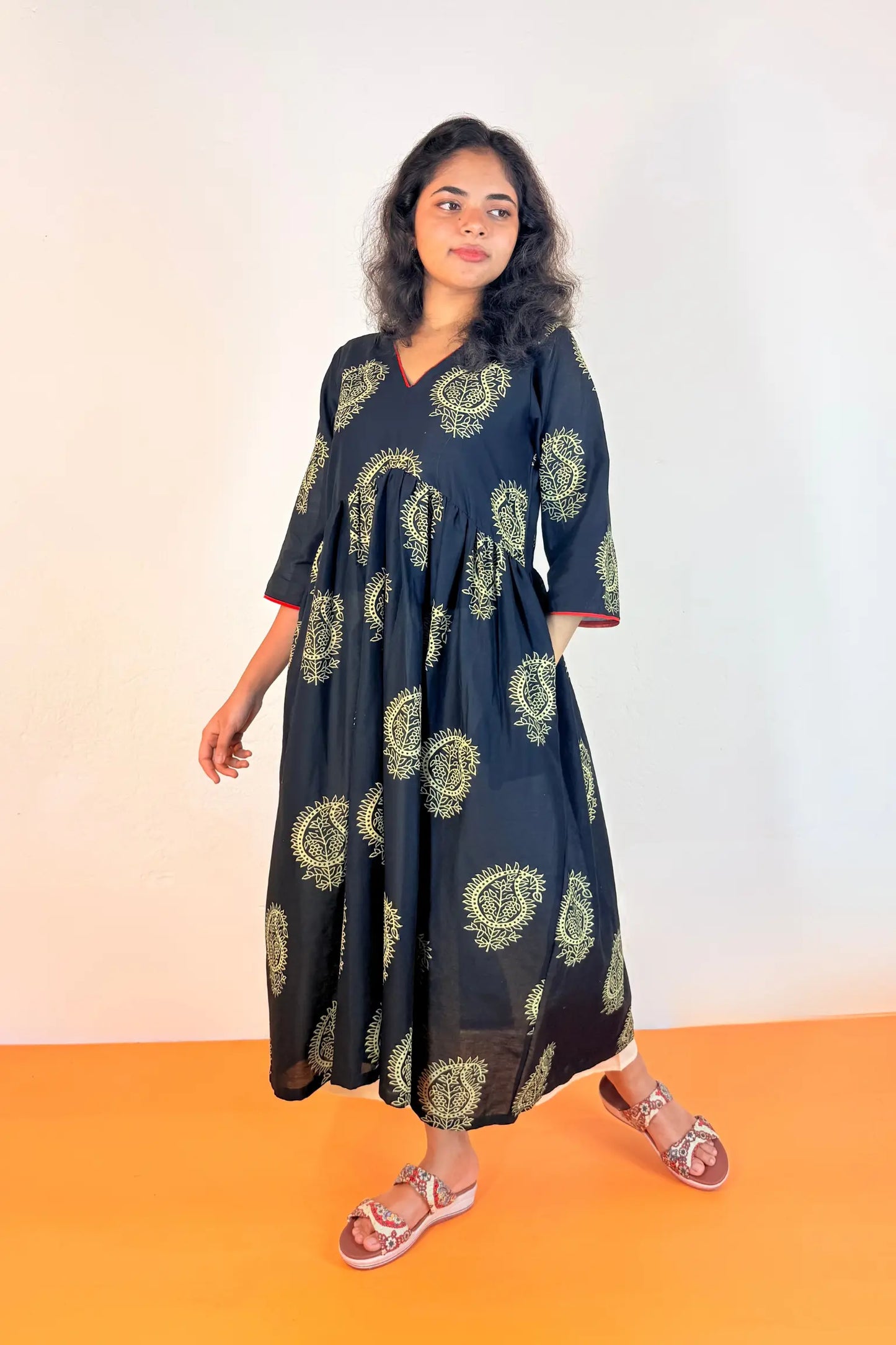 A woman modeling a black V-neck maxi dress with a green and yellow paisley pattern, standing against a white wall with an orange floor.
