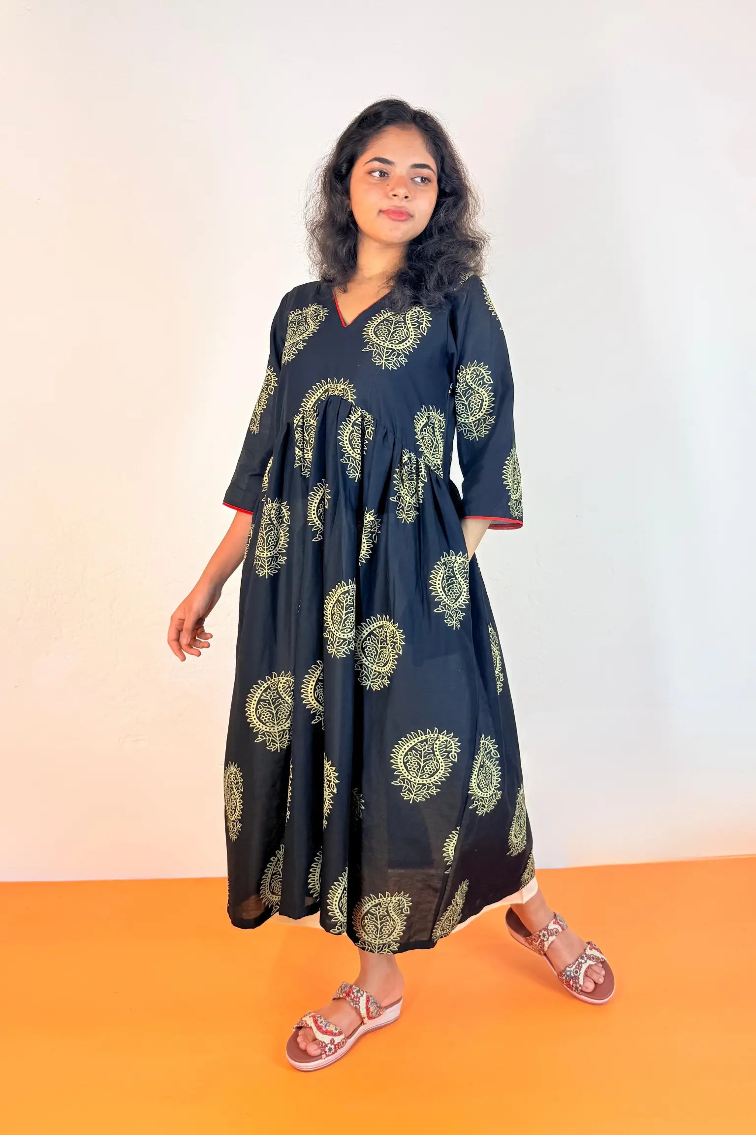A woman modeling a black V-neck maxi dress with a green and yellow paisley pattern, standing against a white wall with an orange floor.