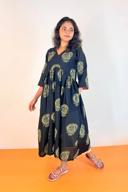 A woman modeling a black V-neck maxi dress with a green and yellow paisley pattern, standing against a white wall with an orange floor.