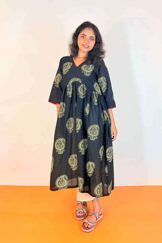 A woman modeling a black cotton maxi dress with olive green circular block prints, standing against a plain white wall with an orange floor.