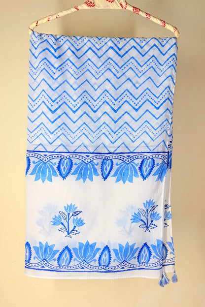 A white fabric towel or scarf with blue patterns, hanging from a patterned wooden hanger against a beige background. The top half features a blue and white chevron pattern, with a blue floral border and blue floral motifs below.