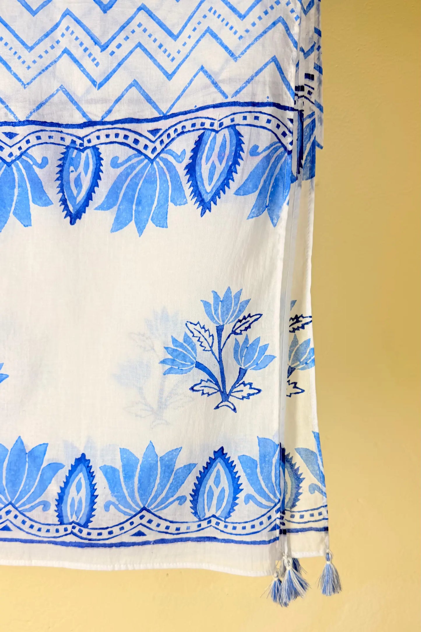 A white fabric scarf or shawl featuring blue block-printed floral and geometric patterns, shown hanging against a solid yellow background.