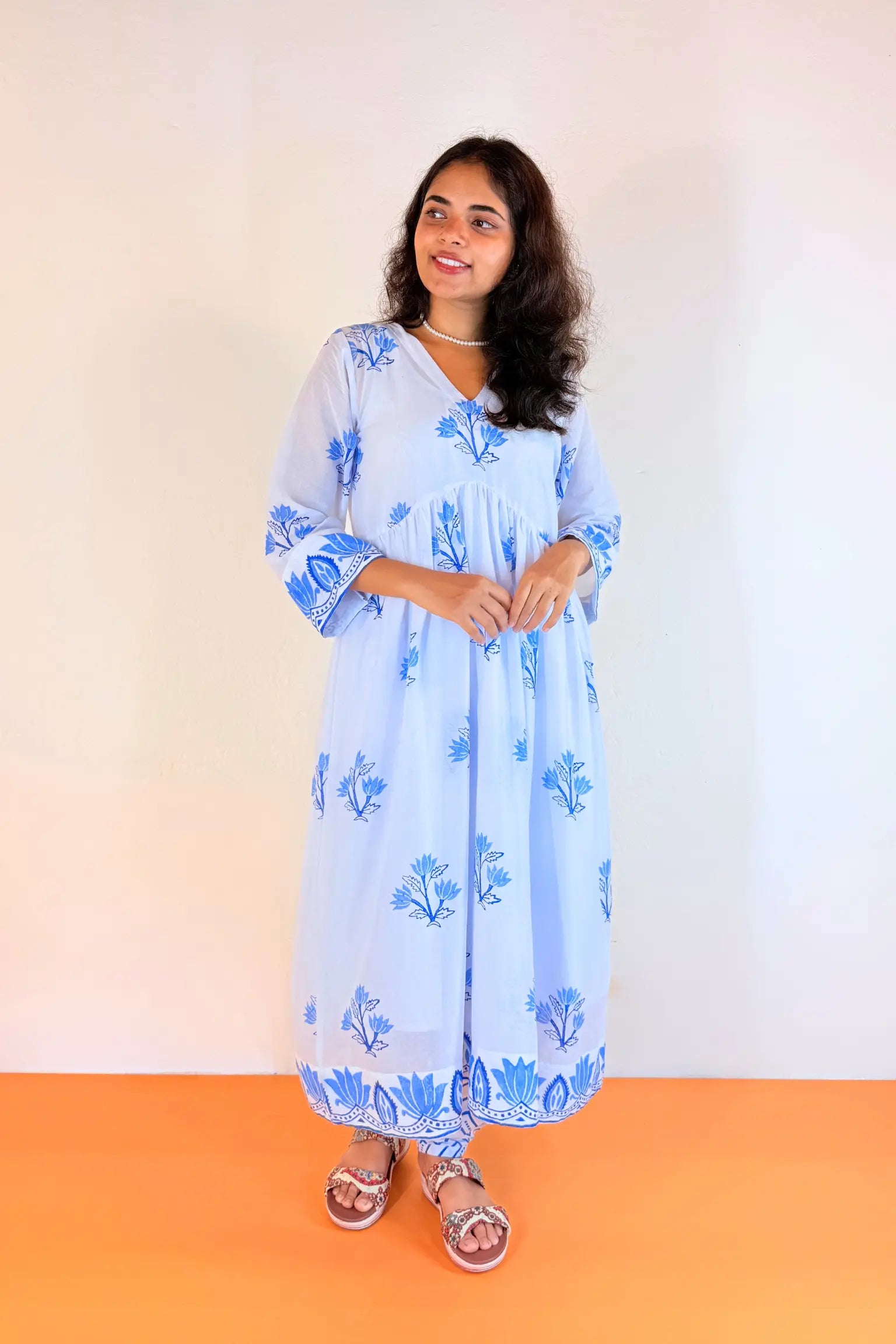 A woman modeling a light blue cotton kaftan dress with blue floral block prints, standing against a plain off-white wall with an orange floor.