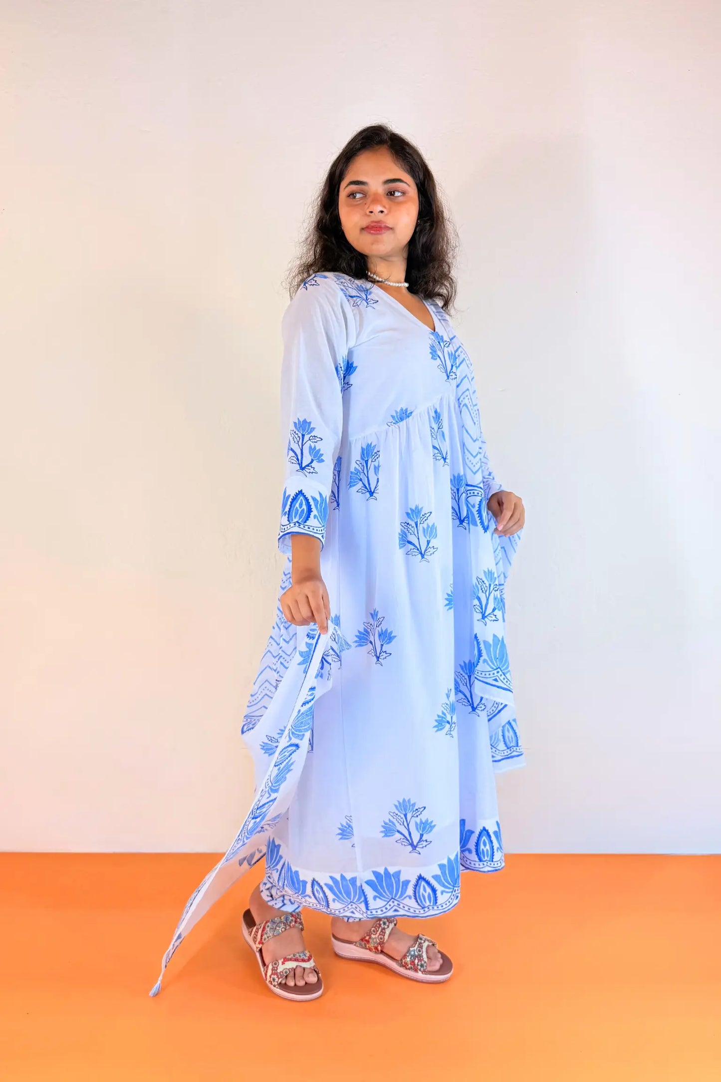 A woman modeling a light blue cotton kurta with blue floral block prints, paired with a matching dupatta and brown sandals, standing against a plain off-white wall and orange floor.