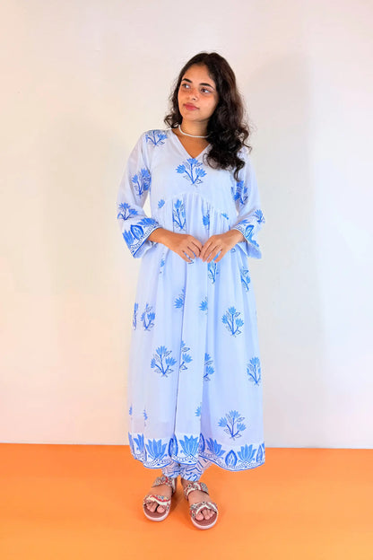 A woman modeling a light blue cotton kaftan dress with blue floral block prints, standing against a plain off-white wall and orange floor.