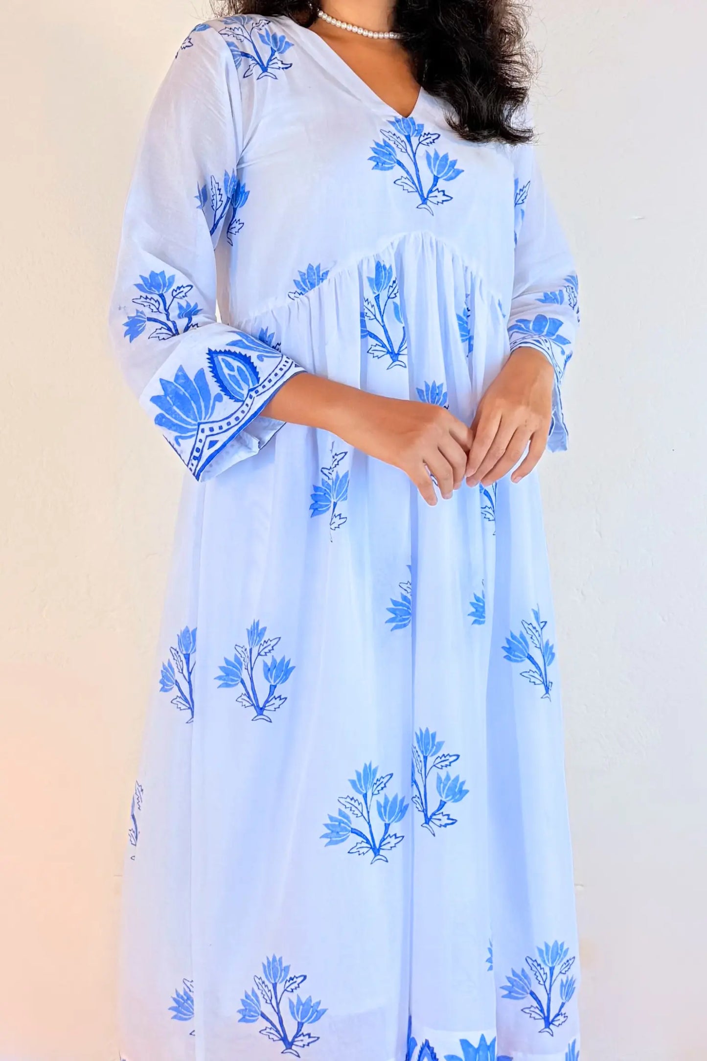 A woman wearing a light blue kaftan dress with blue floral prints and blue patterned cuffs, photographed against a plain light background.