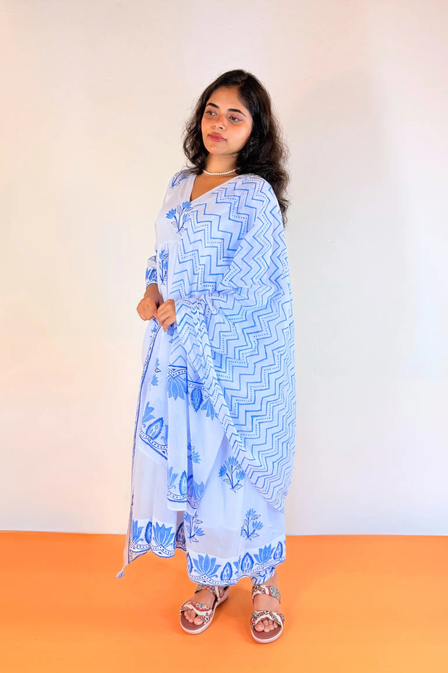 A woman modeling a light blue and white printed traditional outfit with a matching dupatta, standing against a plain off-white wall and orange floor.