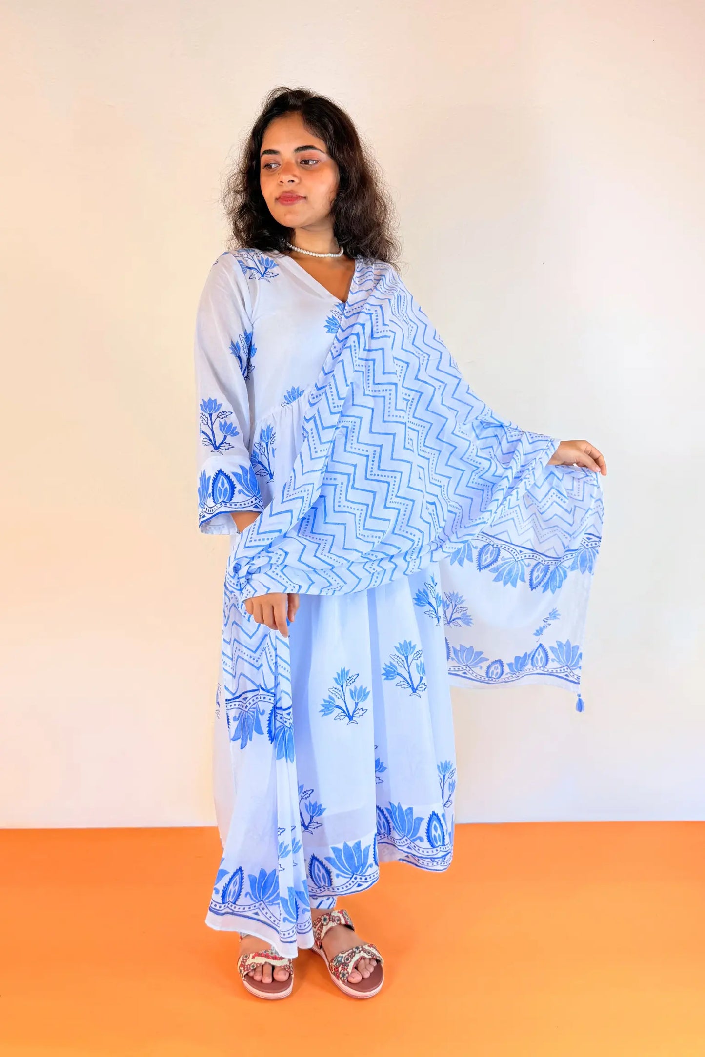 A woman modeling a light blue maxi dress with pink floral embroidery and green leaf details, standing against a plain white wall with an orange floor.