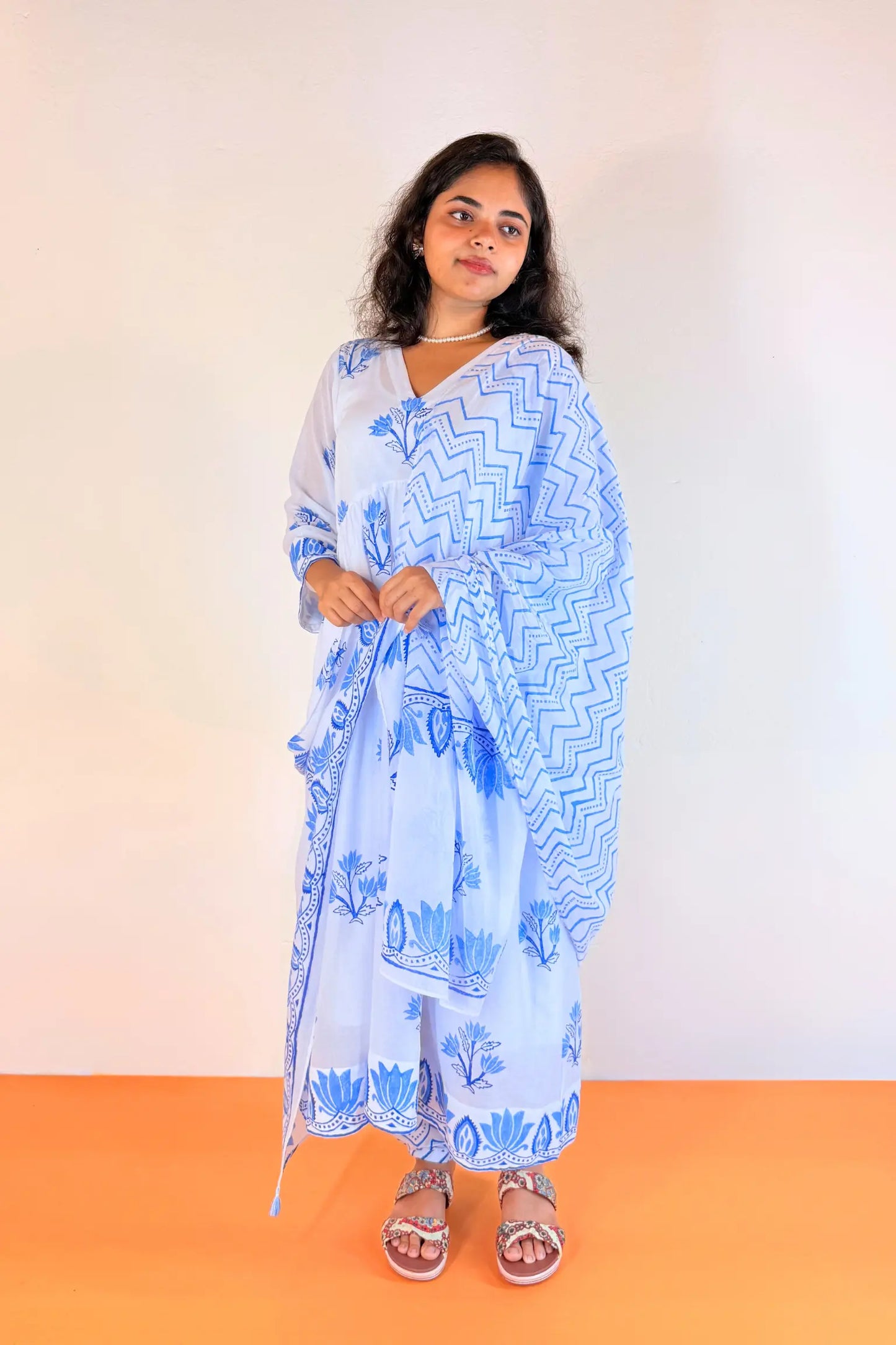 A woman modeling a light blue and white printed traditional outfit with a matching dupatta, standing against a plain off-white wall and orange floor.