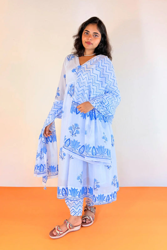 A woman modeling a light blue and white printed traditional outfit with a long kurta, dupatta, and flared pants, paired with brown sandals, set against a plain beige wall and orange floor.