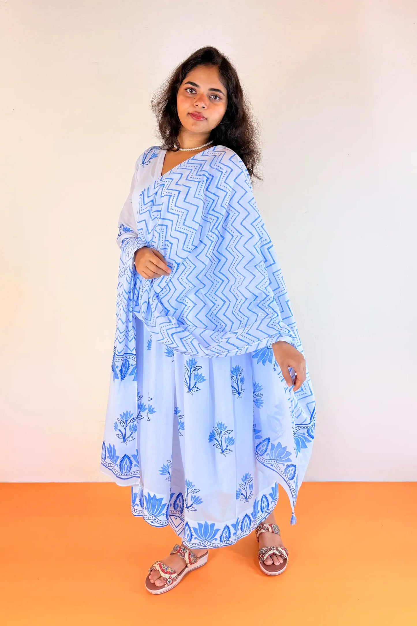 A woman modeling a light blue and white printed traditional outfit consisting of a kurta, matching dupatta, and skirt, paired with brown sandals, set against a plain beige wall and orange floor.