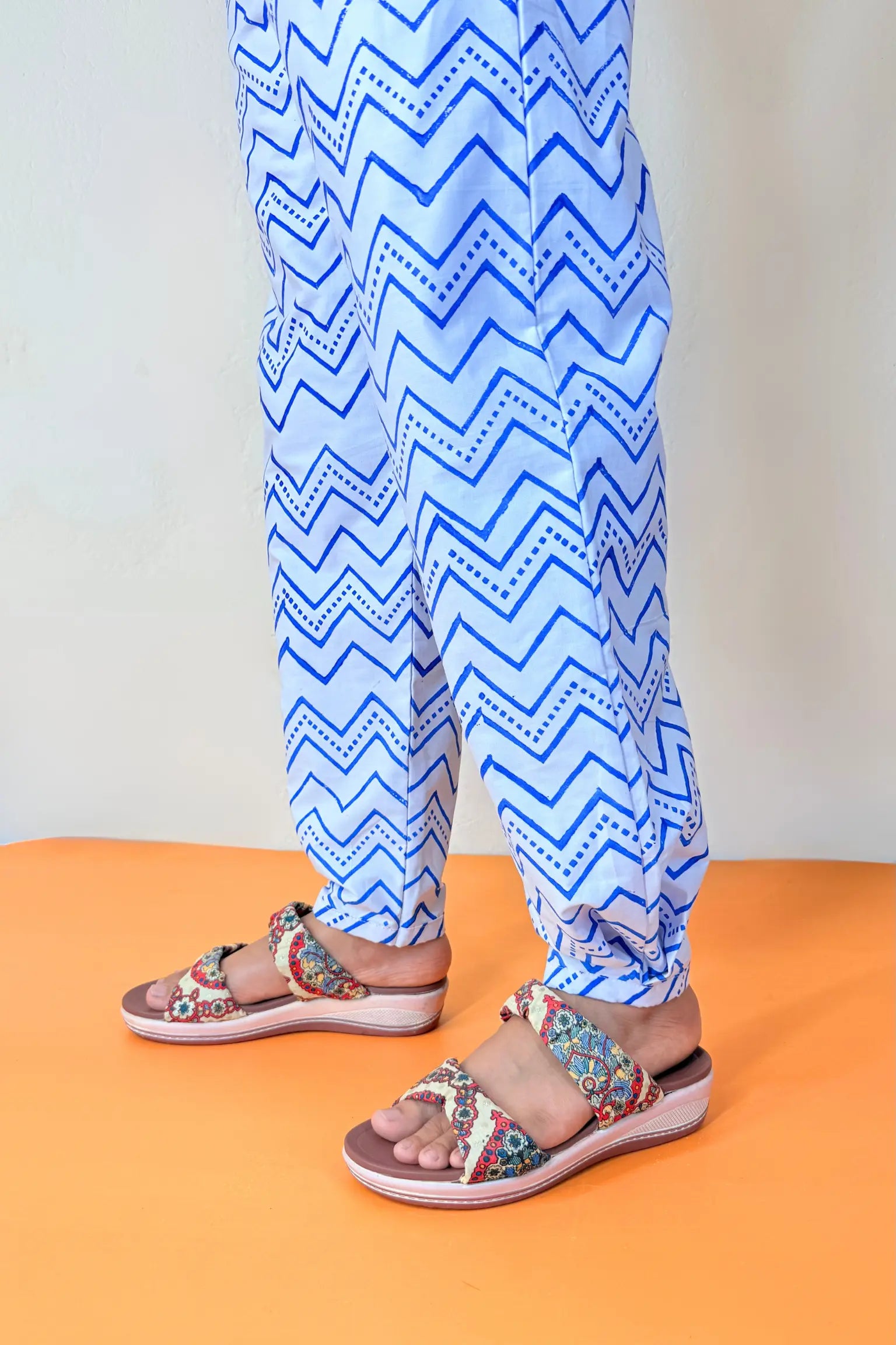 A person wearing blue and white zigzag patterned pants and colorful patterned flat sandals, standing against a light wall with an orange floor.