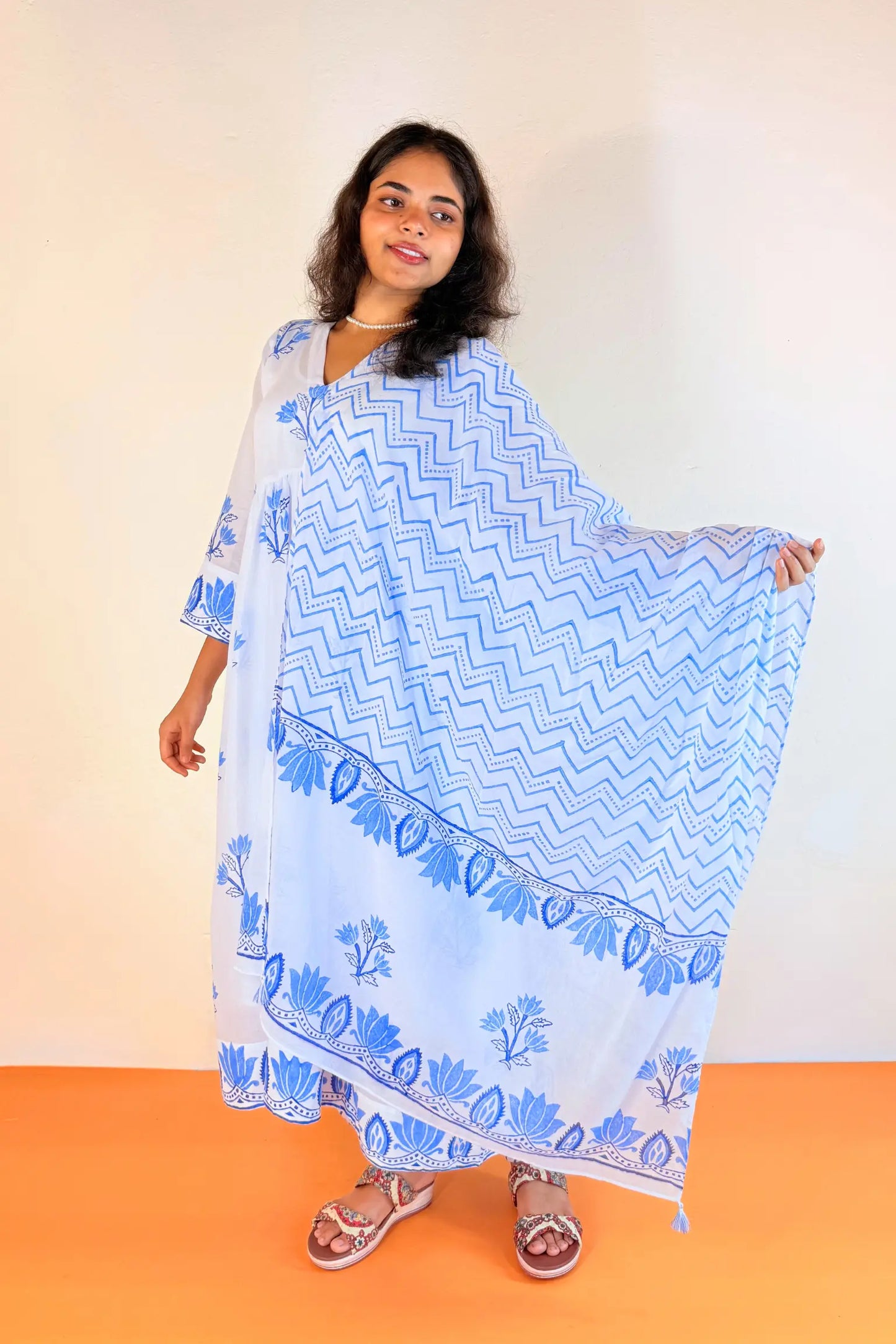 A woman modeling a light blue and white printed kaftan dress, holding out the side of the garment to show its wide patterned fabric. She is standing against a plain beige wall with an orange floor.