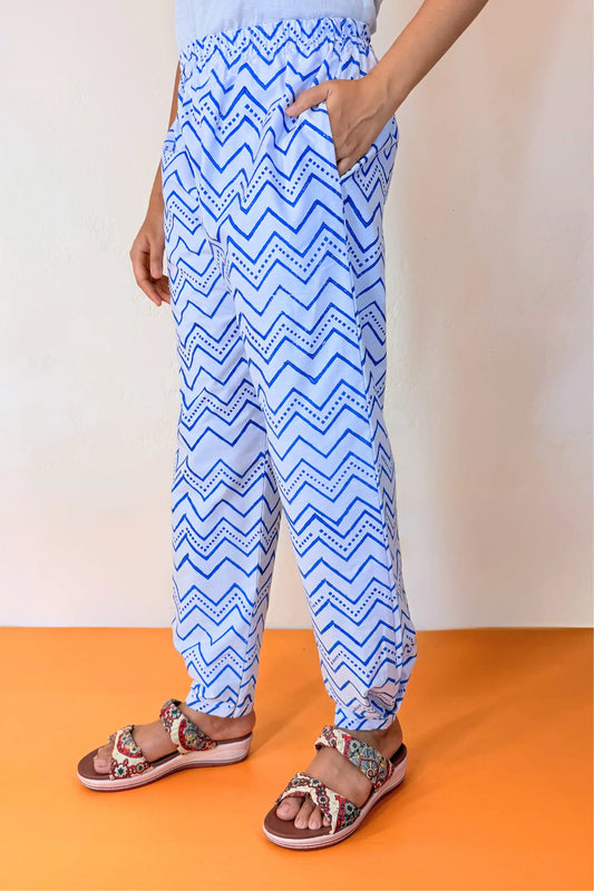 A person wearing loose blue and white zigzag-patterned pants with side pockets, paired with patterned sandals, standing against a plain light wall and orange floor.