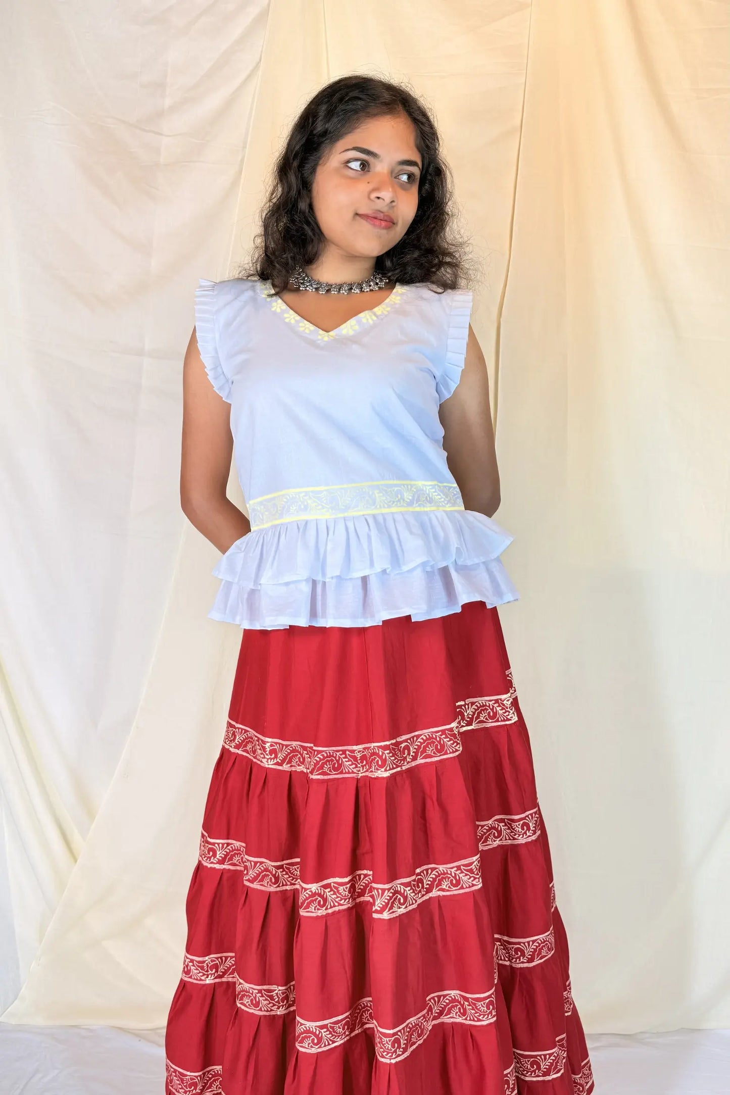 Woman wearing a white top with ruffled layers and a red skirt with white patterns against a plain background
