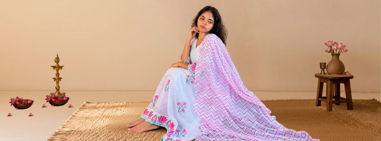 A woman sits on a woven rug in a warm beige room, wearing a white traditional outfit with pink floral embroidery and a light pink patterned dupatta.