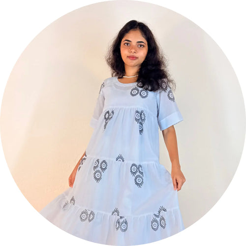 A woman wearing a light blue tiered dress with dark gray floral embroidery, standing against a plain light background inside a circular frame.
