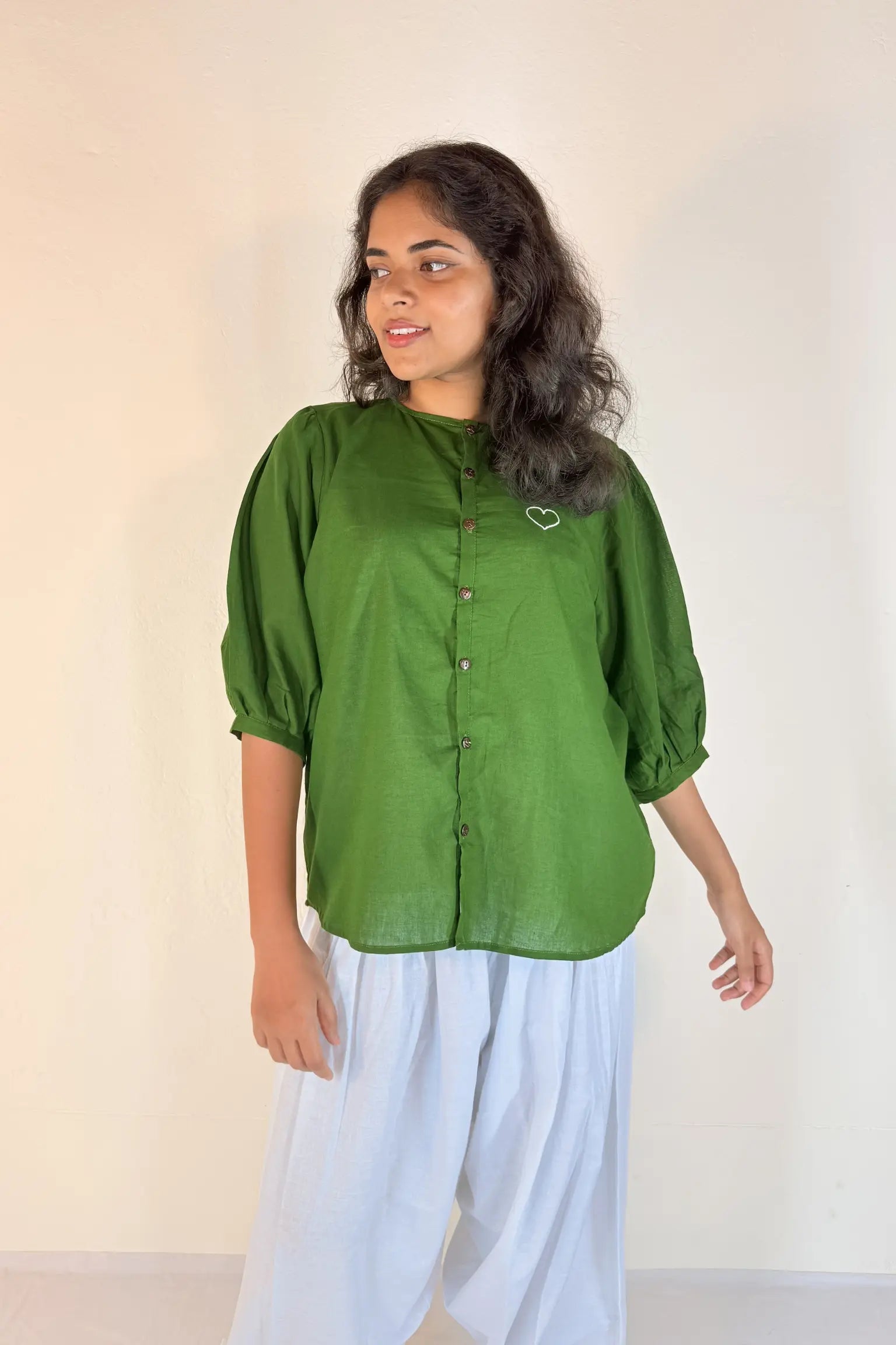 Woman wearing a green blouse with rolled-up sleeves and white pants against a plain background