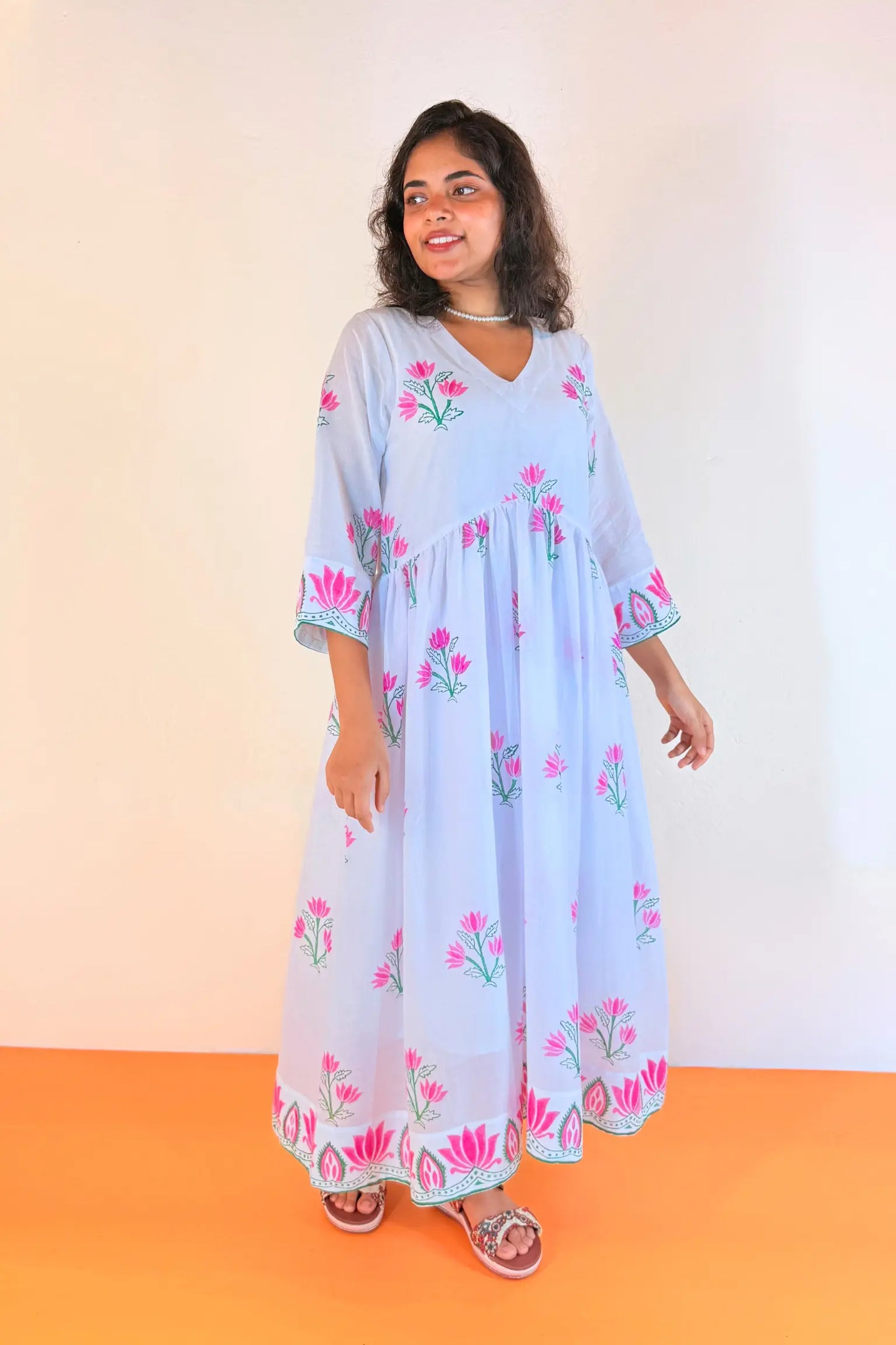 A woman modeling a light blue cotton kaftan dress with pink floral embroidery, standing against a plain off-white wall with an orange floor.