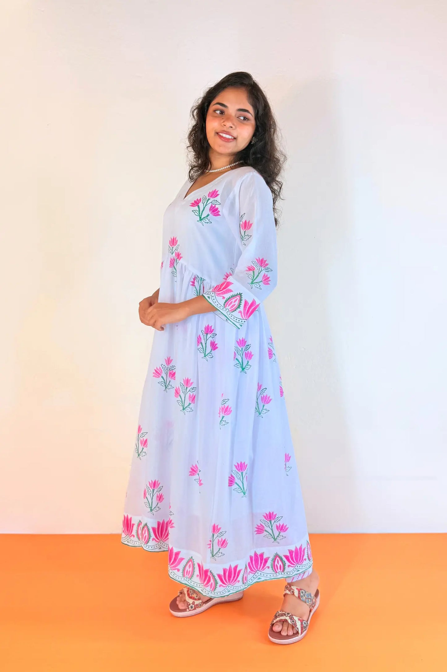 A woman modeling a light blue maxi dress with pink floral embroidery and green leaf details, standing against a plain white wall with an orange floor.