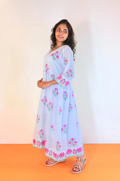 A woman modeling a light blue maxi dress with pink floral embroidery and green leaf details, standing against a plain white wall with an orange floor.