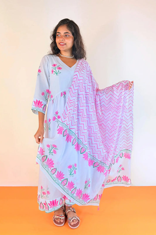 A woman modeling a light blue kaftan dress with pink floral prints and a matching pink and purple chevron dupatta, standing against a plain beige wall and orange floor.