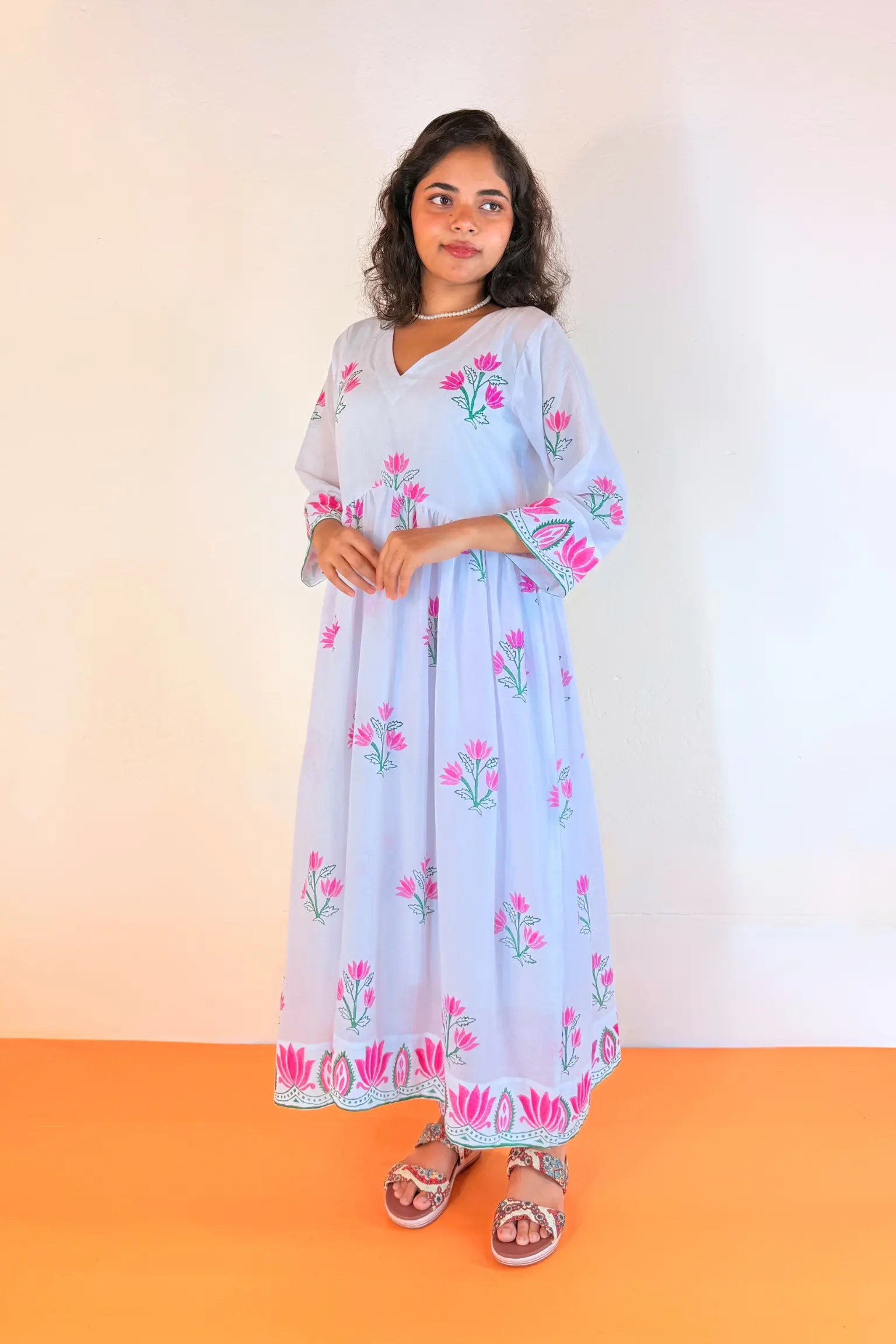 A woman modeling a light blue maxi dress with pink and green floral embroidery, standing against a plain cream wall and orange floor.