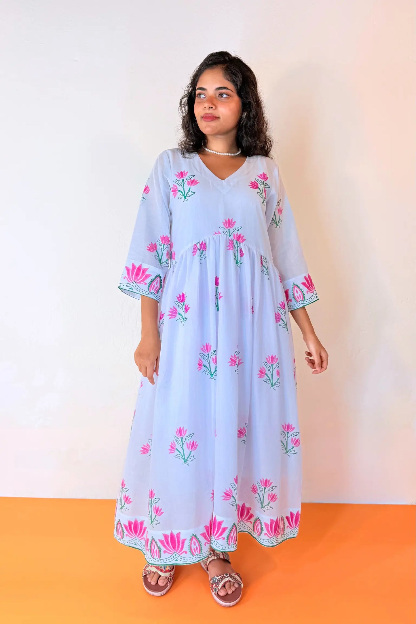 A woman modeling a light blue kaftan dress with pink and green floral embroidery, standing against a white wall with an orange floor.