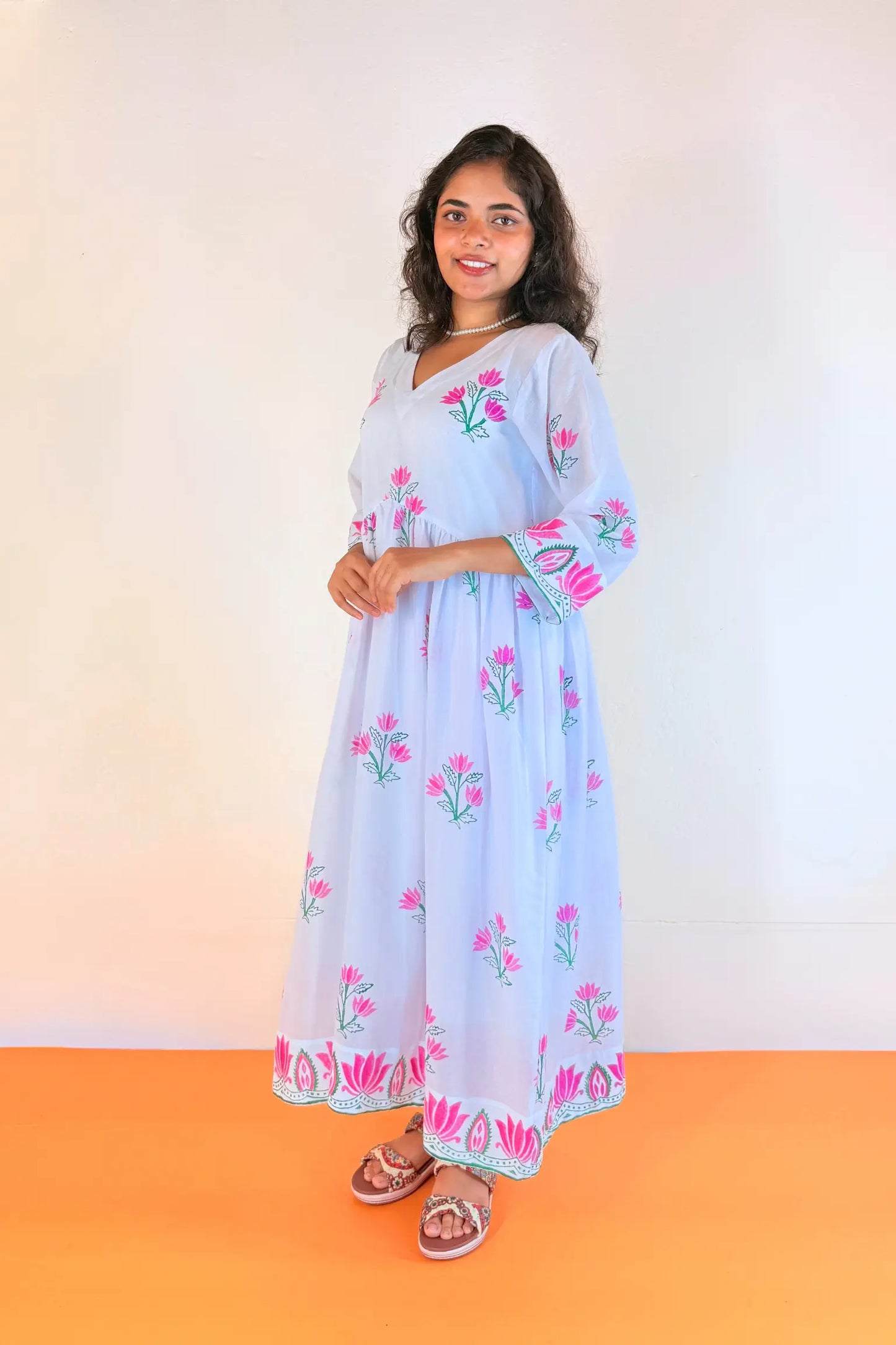 A woman modeling a light blue maxi dress with pink floral embroidery and green leaf details, standing against a plain off-white wall with an orange floor.