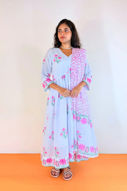 A woman modeling a light blue maxi dress with pink floral prints and a pink patterned dupatta, standing against a plain white wall with an orange floor.