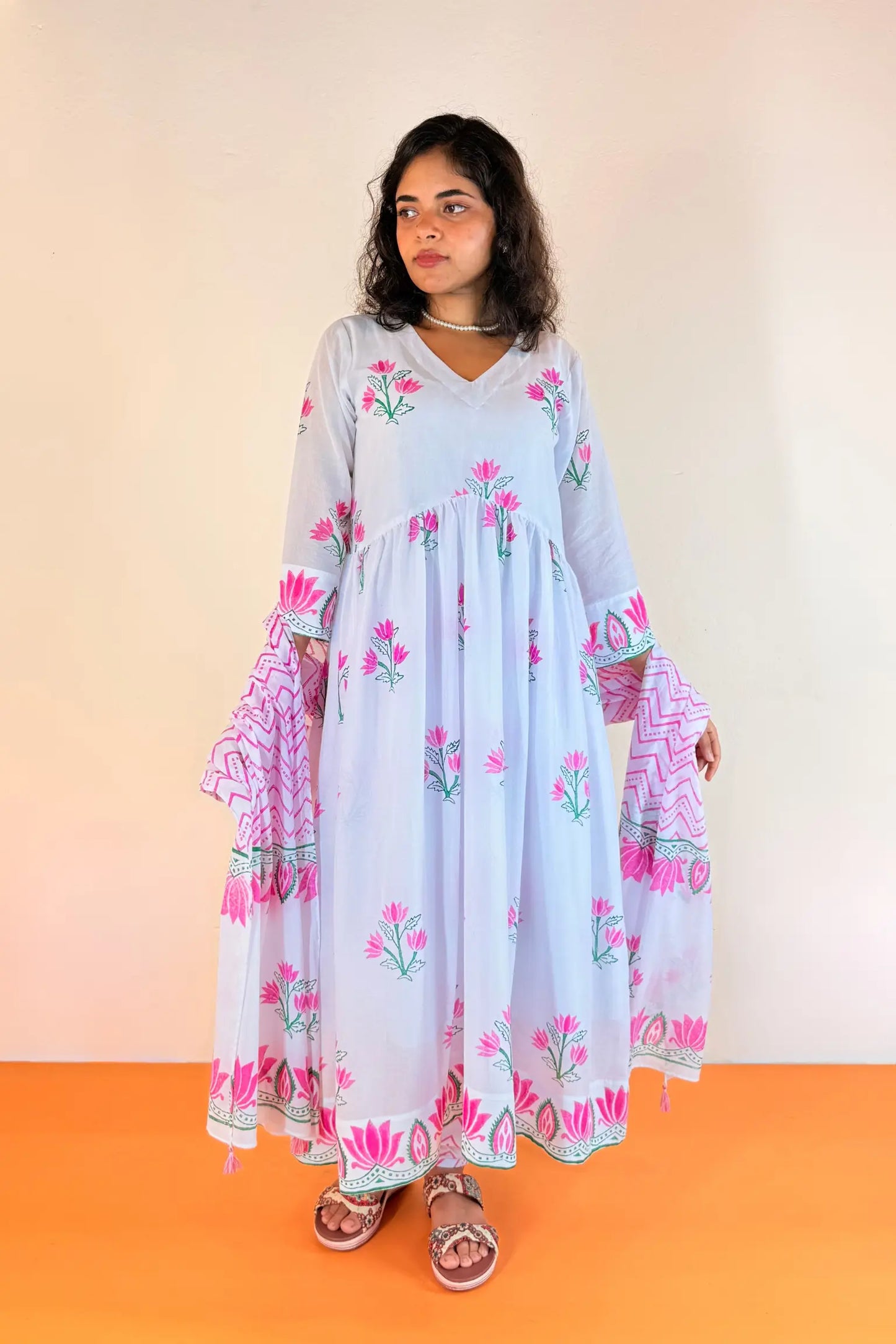 A woman modeling a light blue maxi dress with pink and green floral prints and patterned layered sleeves, standing against a plain beige wall and orange floor.
