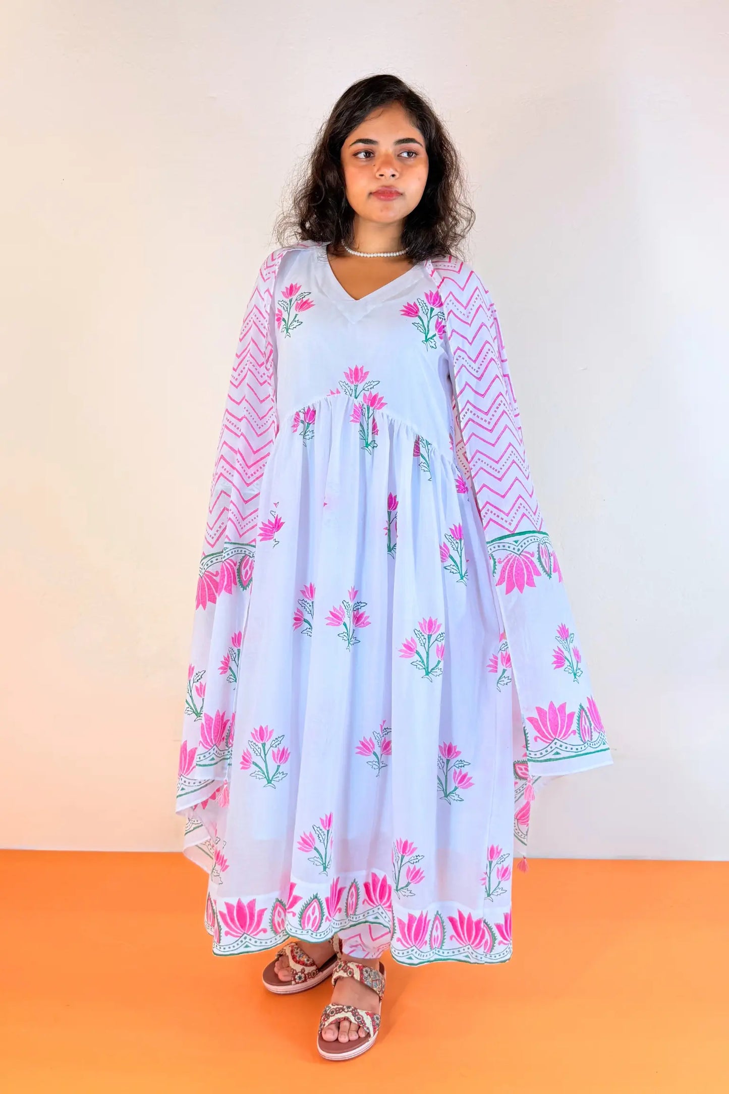 A woman modeling a light blue maxi dress with pink floral prints, pink chevron patterns on the sleeves, and green accents, standing against a plain beige wall and orange floor.