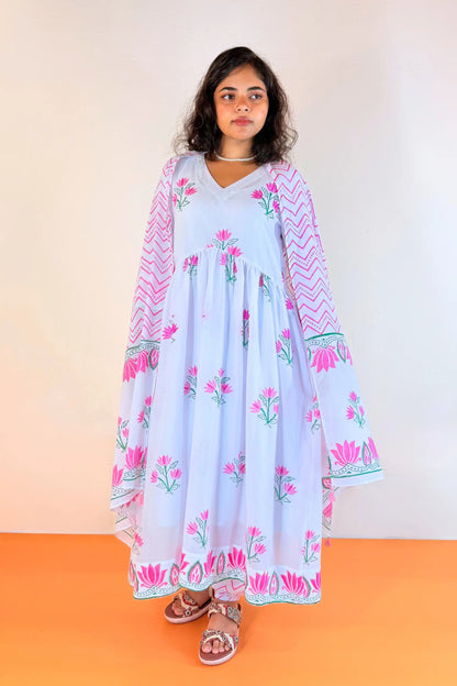 A woman modeling a light blue maxi dress with pink floral prints, pink chevron patterns on the sleeves, and green accents, standing against a plain beige wall and orange floor.