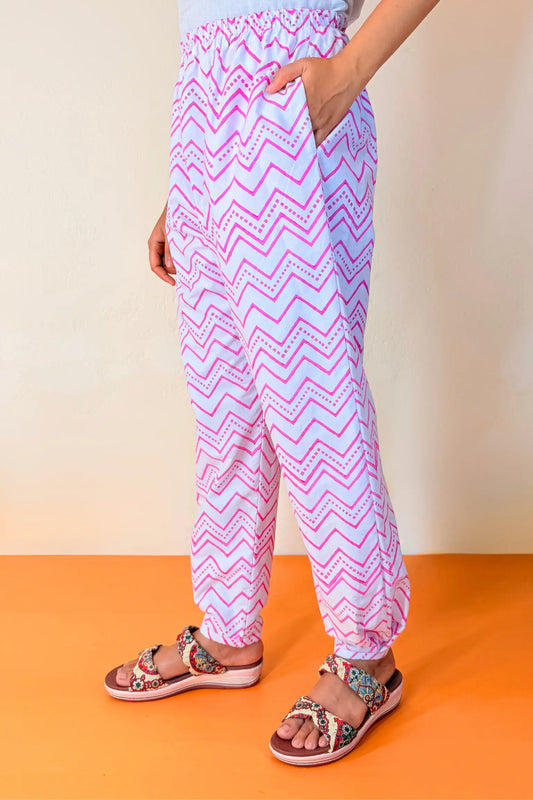 A person wearing loose, high-waisted jogger-style pants with a pink and white chevron pattern, paired with patterned sandals, standing against a beige wall and orange floor.