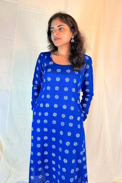 Woman wearing a blue dress with white floral patterns against a white curtain background