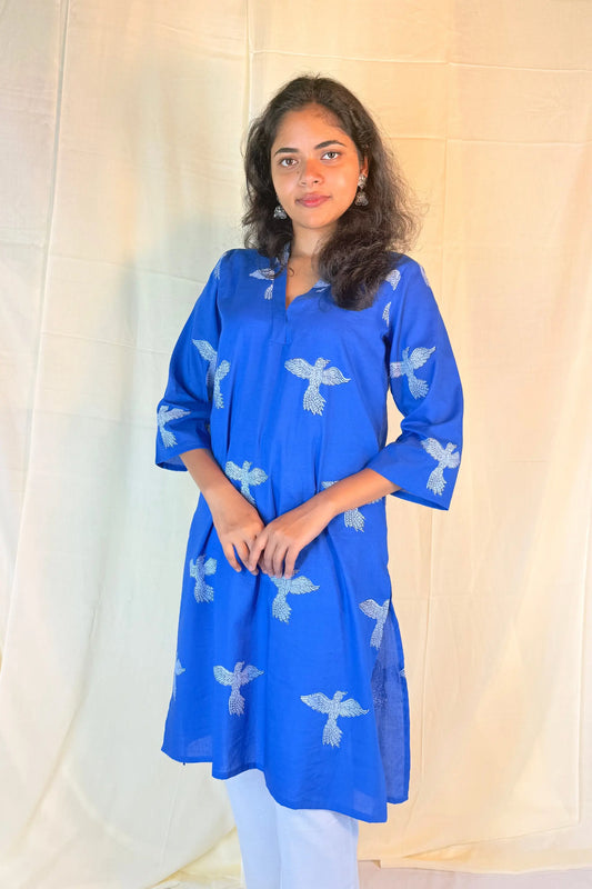 Woman wearing a blue kurta with white patterns against a beige curtain background